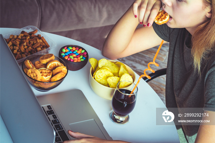 Girl works at a computer and eats fast food. Unhealthy food: chips, crackers, candy, waffles, cola. Junk food, concept.