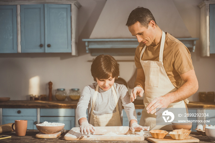 Man and boy standing in kitchen in aprons. Child rolling out batter while father controlling process