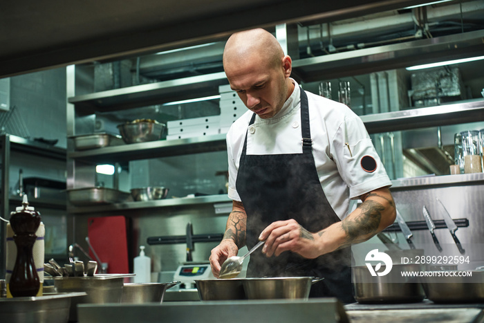 Almost ready. Portrait of handsome chef in black apron and with several tattoos on his arms cooking a dish at his restaurant kitchen