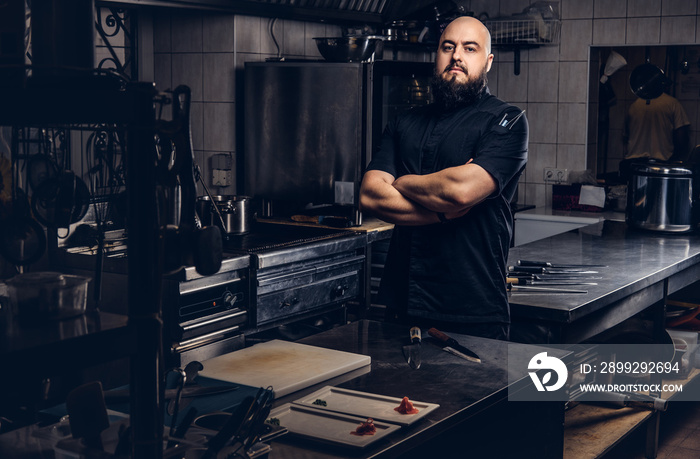 Brutal bearded chef cook in black uniform standing with crossed arms in the kitchen.