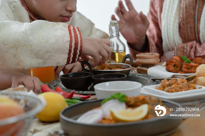 Selective focus on Indian boy hand holding naan bread dipping curry in dish enjoy eating food with hands, wearing traditional clothes, sitting with family at dining table. Indian culture lifestyle