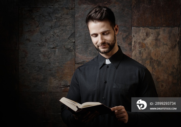 Portrait of handsome young catholic priest reading the prayer book against dark background.