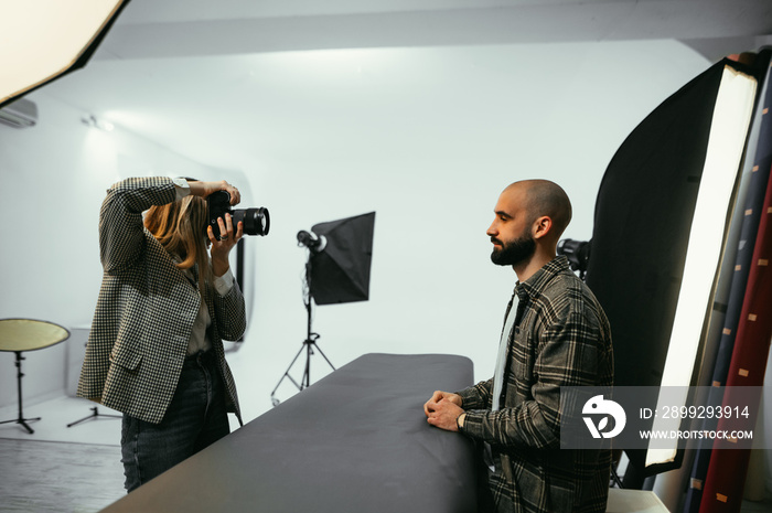 Female photographer works in a photo studio, shoots a sitting bearded man with a pulsed light.