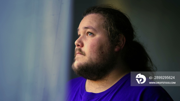 One contemplative casual man standing by window looking out. Calm meditative person at home with thoughtful expression