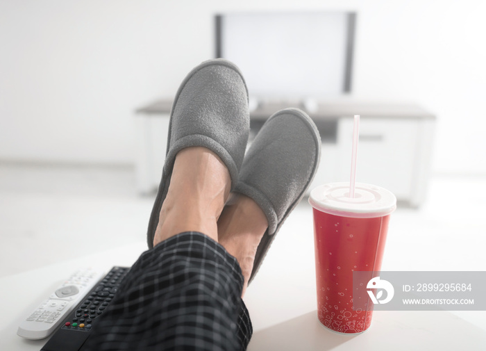 Man drinking soda juice and looking at TV with legs on the table in living room.