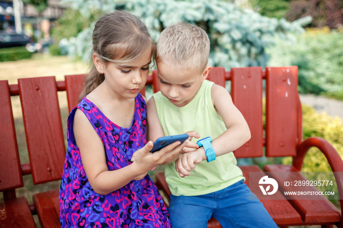 Happy kids sitting on bench and using smartwatches with interest, data synchronization between smartphone and smartwatch, new technology for children, digital education and care, outdoors lifestyle