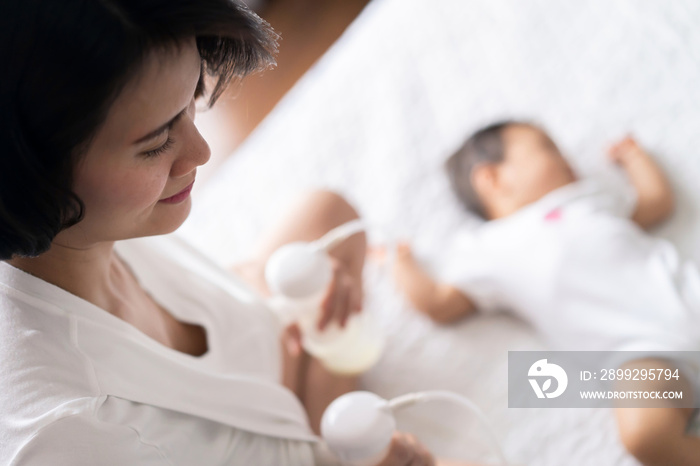 Asian pretty mother pumping breast milk on the bed in bedroom. The baby lying and sleeping on the bed beside her. Mother holding the milk and looking the child with love and smiling. Pump milk concept