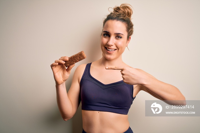 Young beautiful blonde sportswoman wearing sportswear holding energetic protein bar very happy pointing with hand and finger