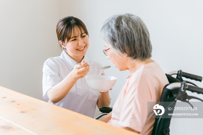 老人ホーム・家で食事する笑顔の高齢者女性と食事介助する管理栄養士・介護士・ヘルパーのアジア人女性