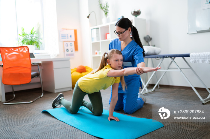 Long-haired therapist in clear glasses assisting young patient