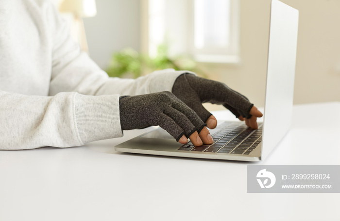 Man with rheumatoid arthritis types text on laptop wearing special compression gloves. Side view close up of hands of young man wearing gray medical cloth gloves struggling with sharp pain his hands