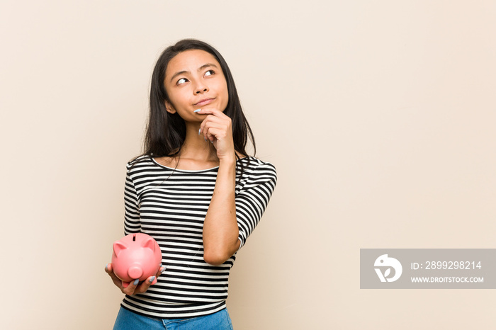 Young asian woman holding a piggy bank looking sideways with doubtful and skeptical expression.