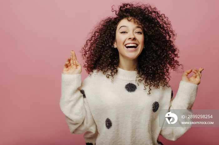 Happy young caucasian woman smiles broadly rejoicing at good news on pink background. Brunette with curly hair wears winter clothes. Emotional response to happiness, concept