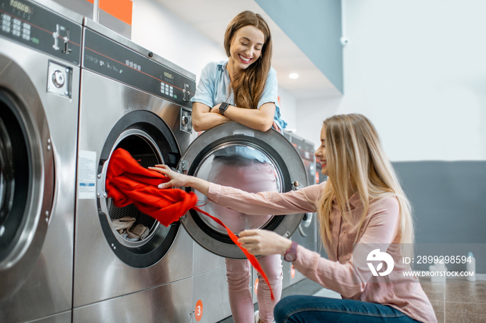 Two cheerful girlfriends having fun loading clothes into the washing machine in the self-service laundry