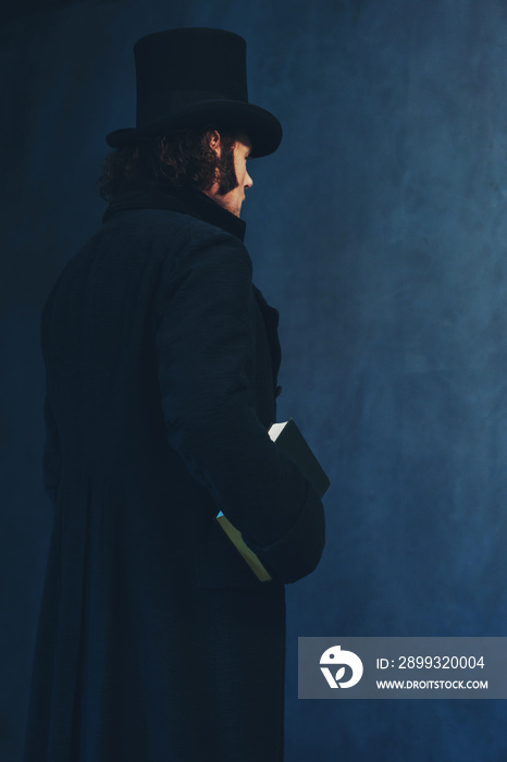 Edwardian man in long black coat and hat holding book standing towards grey wall.