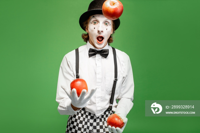 Pantomime with white facial makeup juggling with apples on the green background in the studio