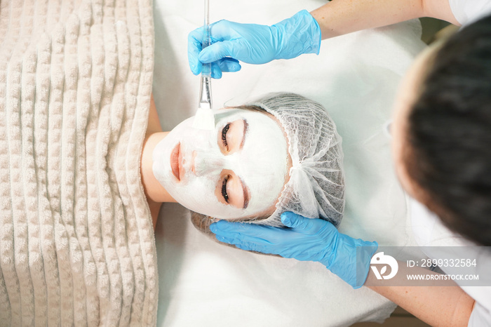 Cosmetologist doing cosmetic face mask treatment for the pretty woman.