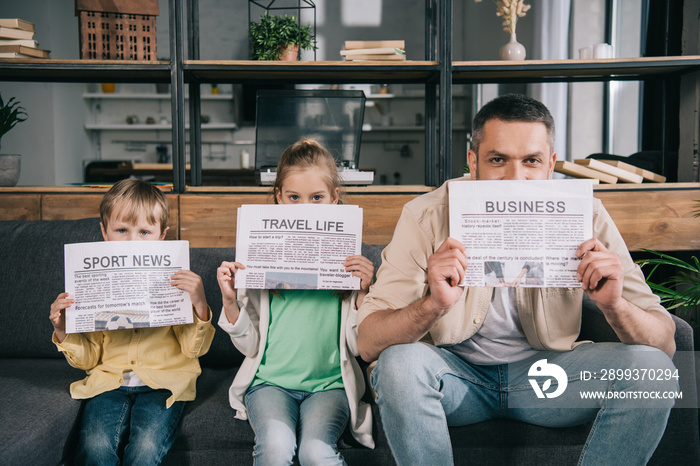 cheerful father and kids holding newspapers while sitting on sofa at home