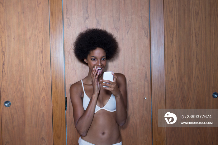 Happy young woman with afro in bra using smart phone at closet