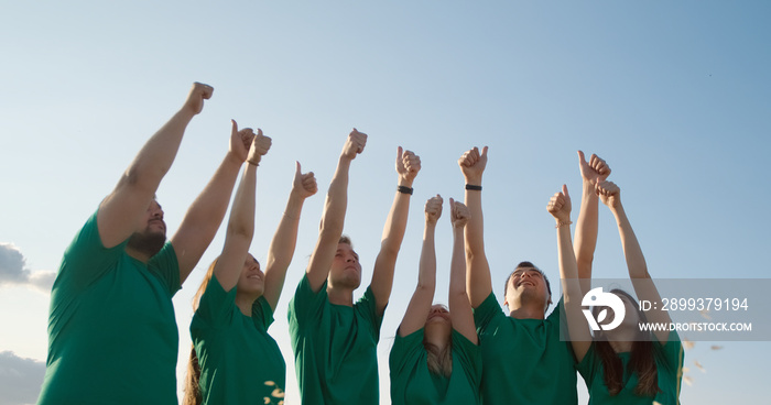 Group of happy young people in green T-shirts raise their hands up and show thumbs up, on the backgr