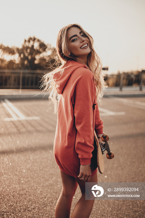 Positive girl walking on city street with skateboard in urban red hoodie. Happy attractive young wom