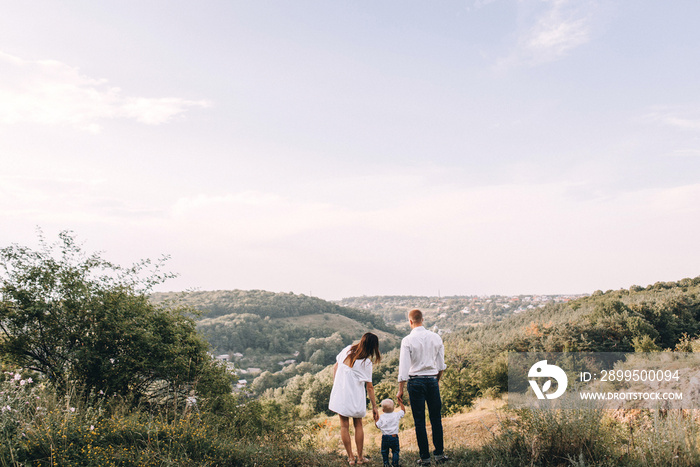 Walk beautiful young family in white clothes with a young son blond in mountainous areas with tall grass at sunset. Mother keeps son in her arms, hugging. family - this is happiness