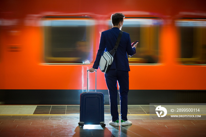 Young stylish handsome man in suit with suitcase standing on metro station holding smart phone in hand, scrolling and texting, smiling and laughing.  Orange train passing by. Futuristic bright subway