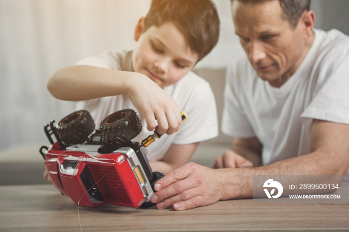 Interested boy is repairing toy by instrument with help of his dad. Focus on small red car