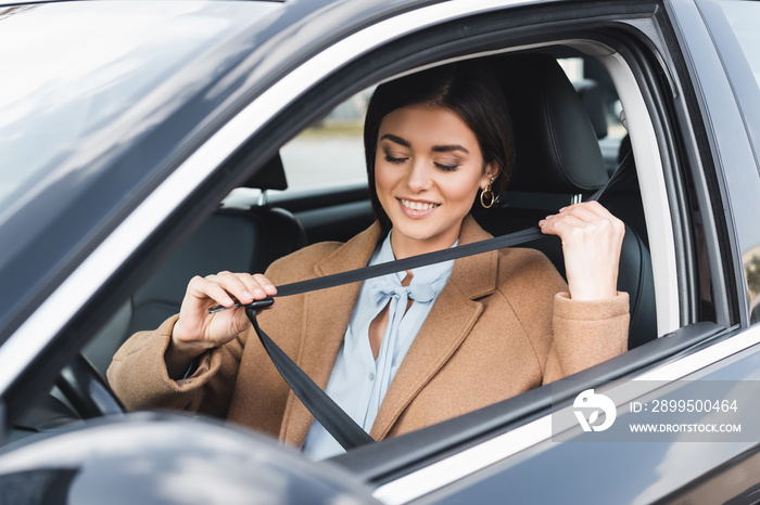 woman in autumn outfit putting on safety belt while sitting in car on blurred foreground