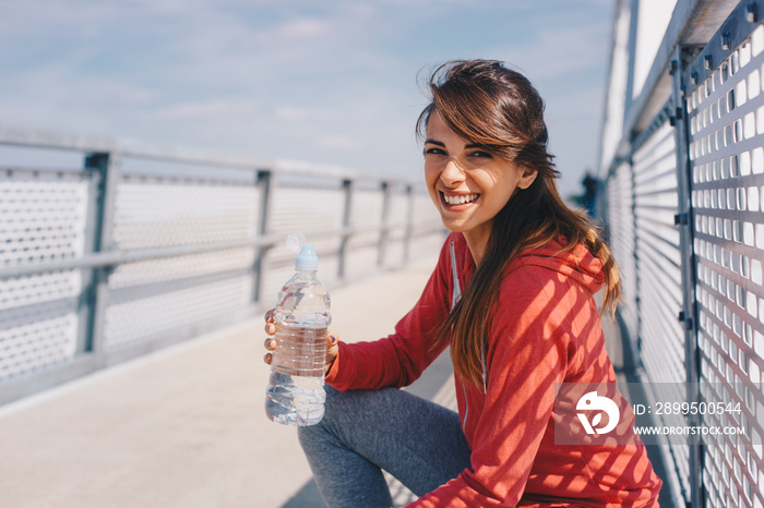 A woman crouching on a bridge after jogging. She is holding a bottle of water and smiles, looking at the camera.
