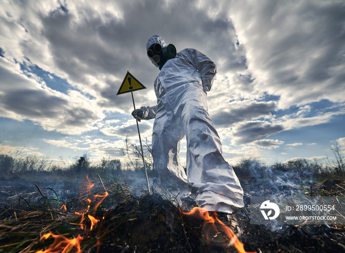 Fireman ecologist fighting fire in field. Man in protective radiation suit and gas mask near burning grass with smoke, holding warning sign with exclamation mark. Natural disaster concept.