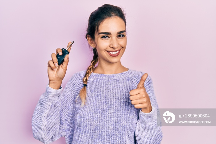 Young hispanic girl holding key of new car smiling happy and positive, thumb up doing excellent and approval sign