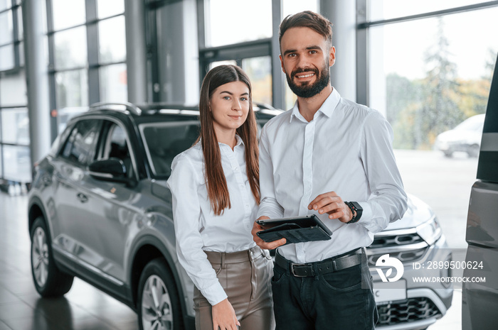 Standing together and smiling. Young man in white clothes is in the car dealership with woman