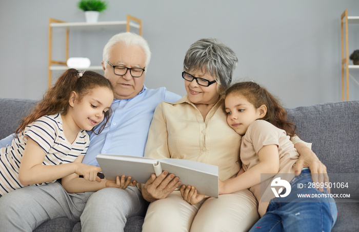 Happy grandparents and grandchildren spending time at home together. Grandma, grandpa and grandkids sitting on the sofa in the living room, reading a book or looking through a family photo album