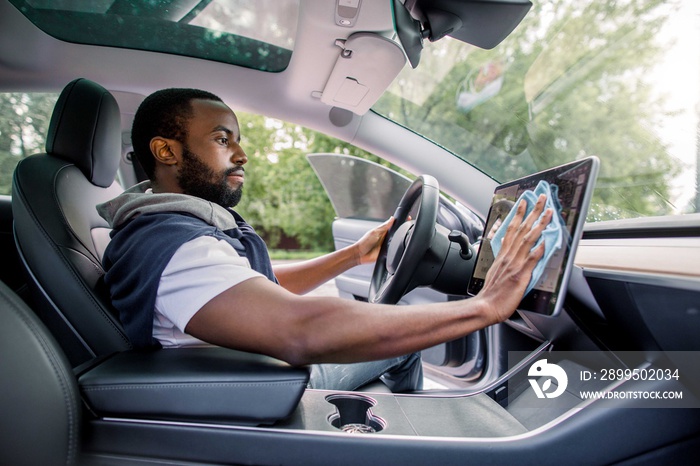 Cleaning time, car detailing concept. Portrait of young handsome African man with beard sitting inside his modern self-steering car and wiping dust from touchscreen display with microfiber cloth