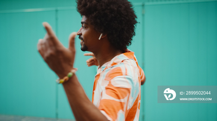 Close-up of cheerful young African American man wearing shirt listening to music in wireless headphones and dancing on light blue wall background. Lifestyle concept.