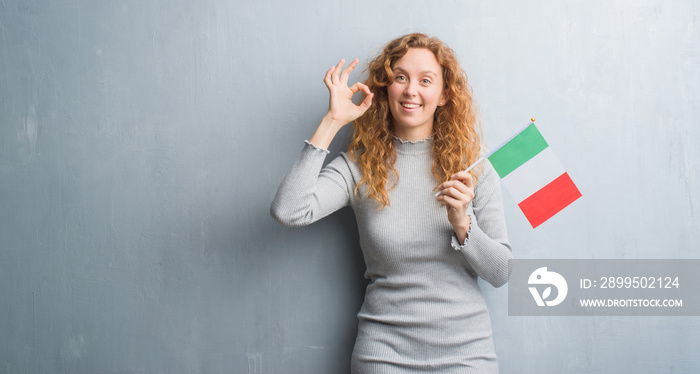 Young redhead woman over grey grunge wall holding flag of Italy doing ok sign with fingers, excellent symbol