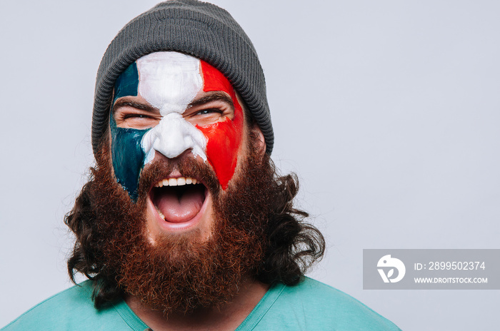 Face of young happy bearded man painted with flag of France. Football or soccer team fan, sport event, faceart and patriotism concept. Studio shot at gray background, copy space