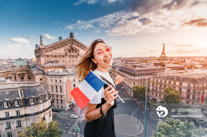 Happy asian woman with french flag and backpack standing on a roof top and enjoys great view over Parisian skyline at sunset. Travel and education in France