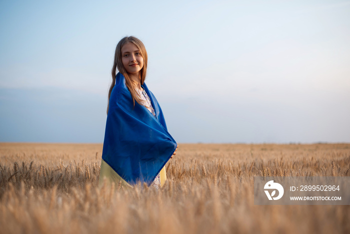 Smiling girl wrapped in ukrainian flag in the boundless field of rye at sunset. Patriotic ukrainian girl