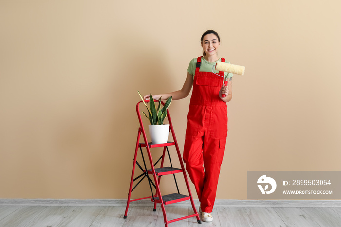 Young woman with paint roller, ladder and houseplant near beige wall