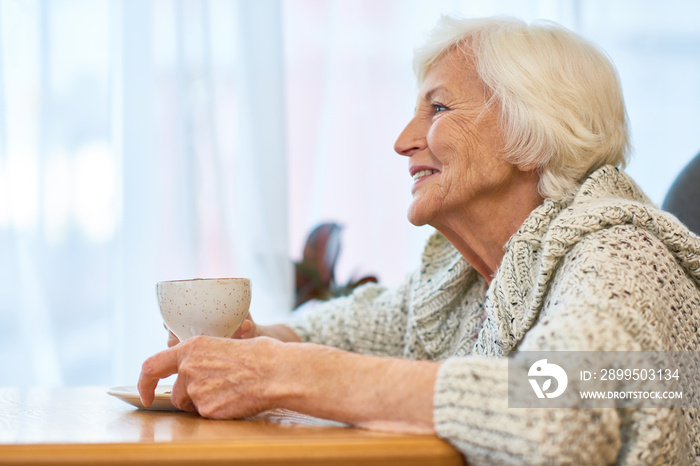 Profile view of cheerful aged woman wearing knitted sweater enjoying fragrant coffee while sitting at table of cozy small cafe