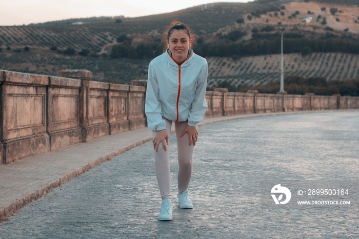 Niña joven de pelo castaño oscuro ejercitándose por la mañana junto a las montañas flexionando las rodillas para recorrer en ruta la ciudad y quemar calorías