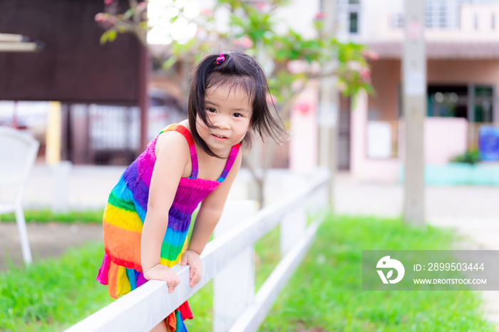 Asian girl wearing a rainbow-colored dress is climbing the white fence of playground to cross shore. Field has green grass. Girl smiled sweetly in summer or spring. 3 year old little cute and bright.