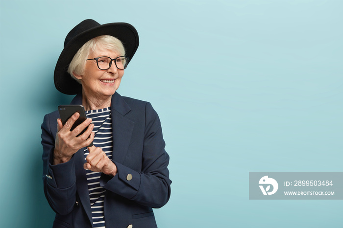 Old attractive woman types text message on cellular, uses new online application, has positive facial expression, looks away, wears stylish headgear and coat, stands over blue studio wall, free space