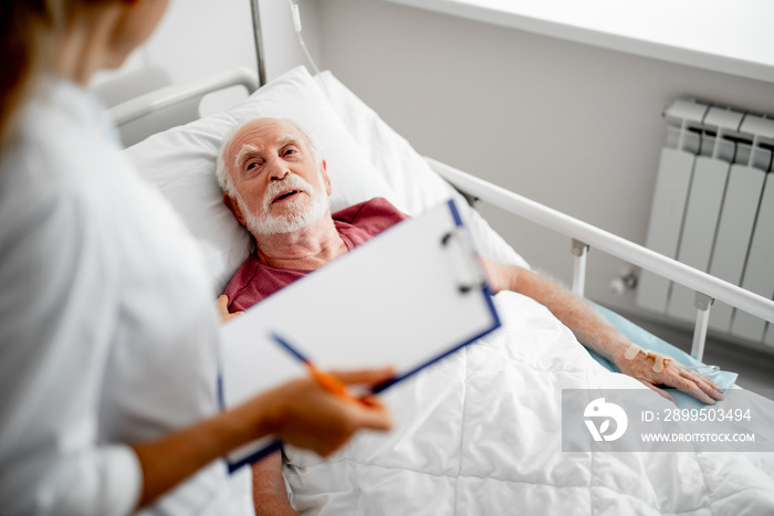 Thank you for your concern. Top view portrait of old man resting in hospital room and chatting with female therapist. Focus on bearded gentleman