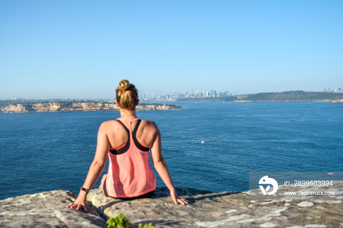 Morning view of a young female hiker sitting on a rock at North Head, a headland in Manly and part of Sydney Harbour National Park in Sydney, New South Wales, Australia. Sydney skyline in background.