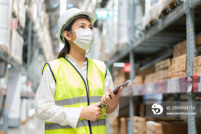 Young asian woman auditor or trainee staff wears mask working during the COVID pandemic in store warehouse shipping industrial. looking up and checks the number of items store by digital tablet.