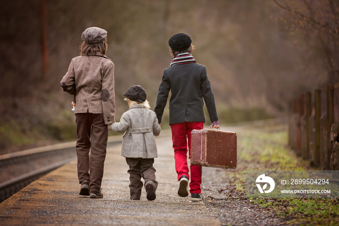 Adorable boys on a railway station, waiting for the train with suitcase and beautiful vintage doll