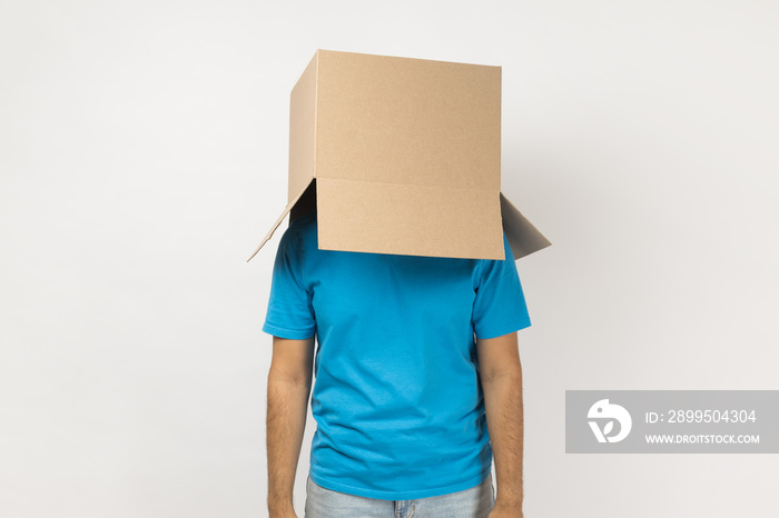 Portrait of unknown anonymous man wearing blue T- shirt standing with cardboard box on his head, having fun, hiding his face in carton parcel. Indoor studio shot isolated on gray background.